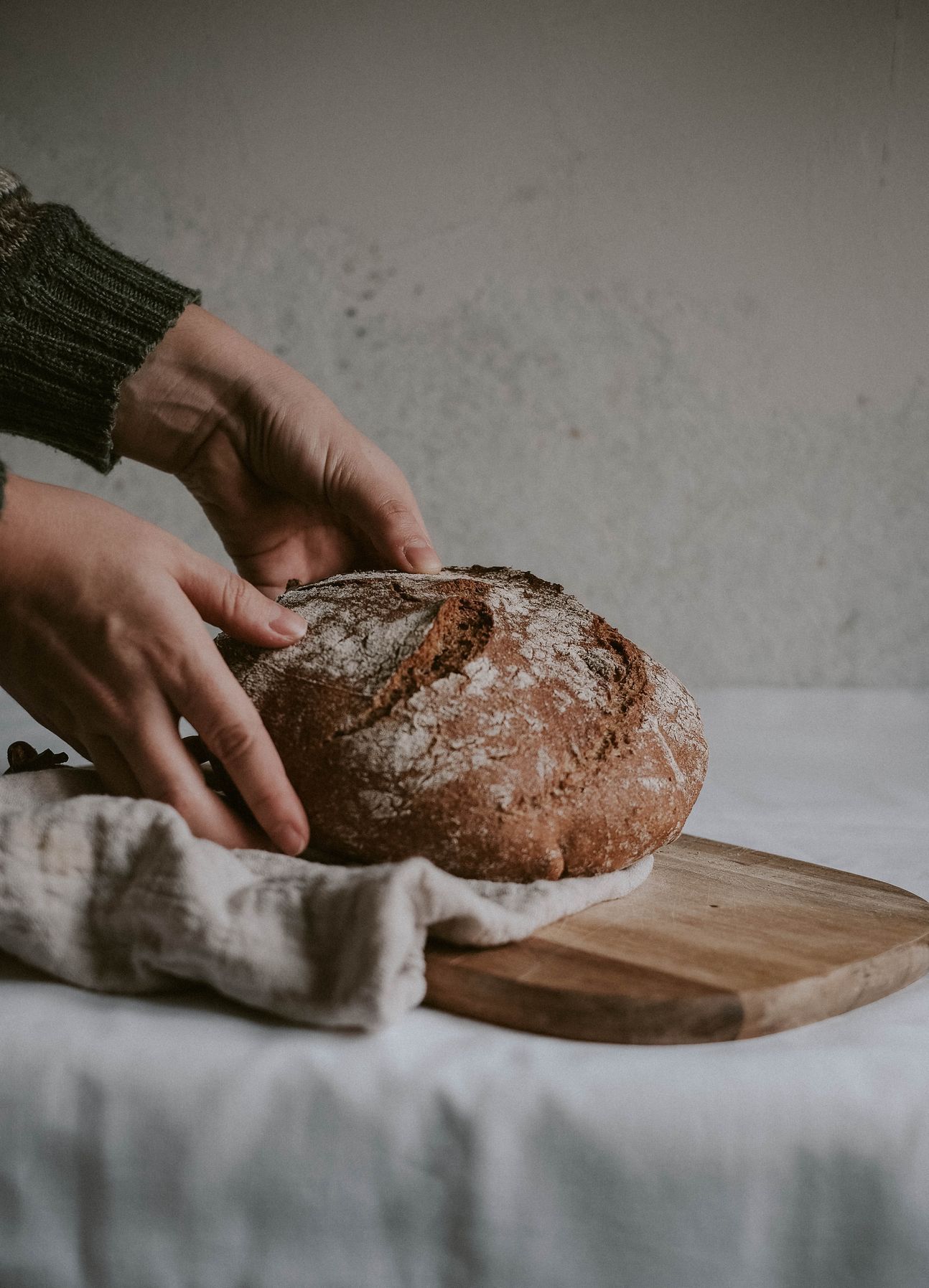 Ein Laib Brot auf einem Holzbrett, umfasst von zwei Händen und ein Tuch.