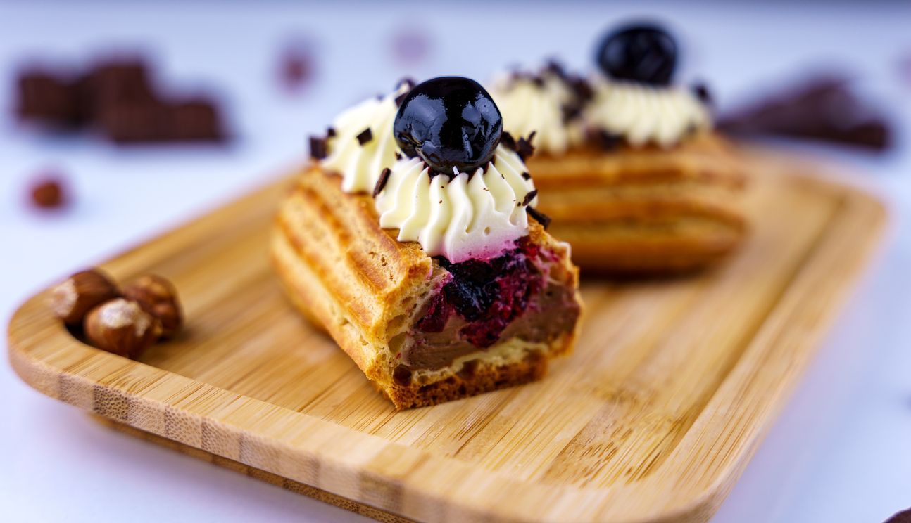 Leckere Eclairs, auf einem Holzbrett angerichtet, mit Haselnüssen dekoriert. Auf den Eclairs Sahne und Heidelbeeren.