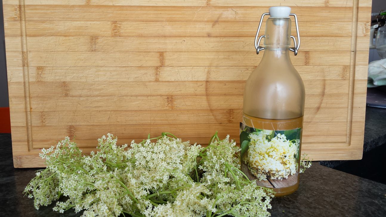 Eine Flasche mit Flüssigkeit vor einer Flechtwand, daneben Holunderblüten.