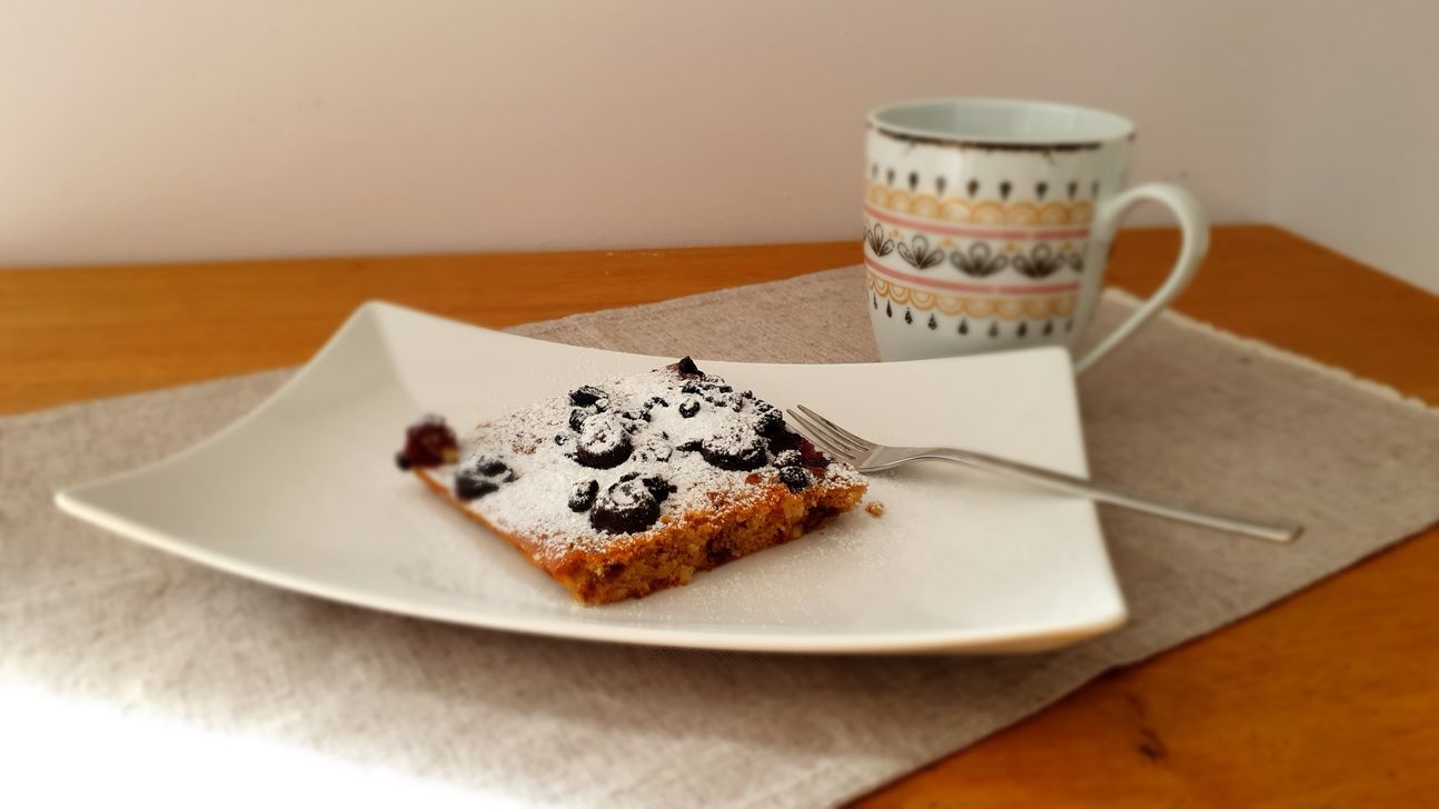 Schokoladen-Nuss-Kuchen mit Beeren und Puderzucker auf einem weißen, rechteckigen Teller und im Hintergrund eine Tasse.